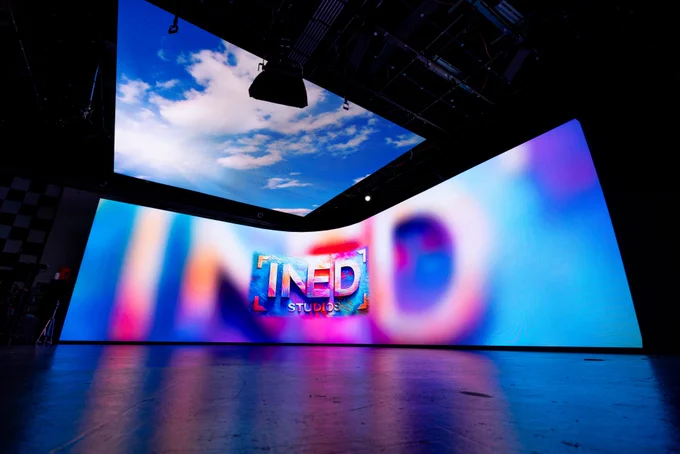 A digital screen in an indoor studio displaying a colorful, abstract image with the words "INED STUDIOS." The ceiling screen shows a clear blue sky with clouds.