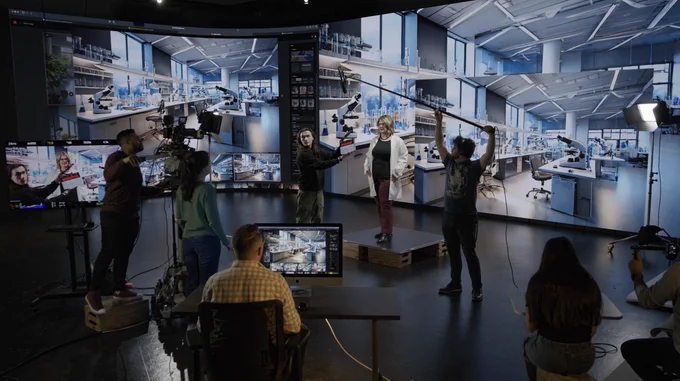 A group of people are on a production set with large digital screens displaying a modern office. Cameras and lighting equipment surround the area, and several individuals handle microphones and cameras.