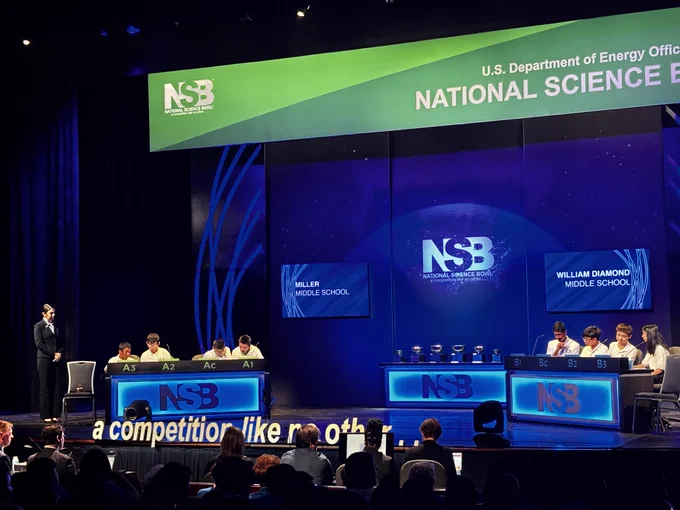 The middle school teams competing. There are four students on each team, standing behind a lit-up podium-like table. Each person has a tag with their designation on it in front of them. There is a banner that says National Science Bowl above them and televisions with the name of their team on it.