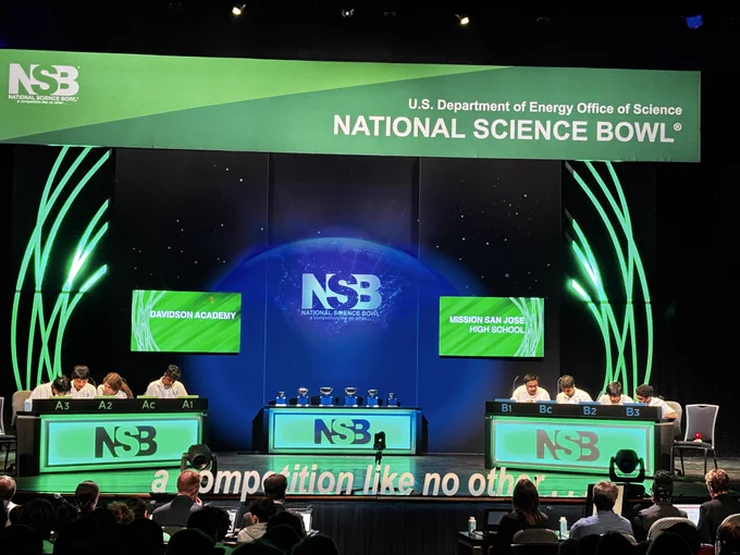 The final two high school teams competing against each other on stage. Each team has four students sitting behind a table labeled NSB with labels indicating their designation. A banner overhead says "National Science Bowl" and televisions behind them say the name of their schools.