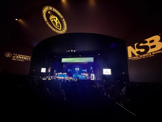 The middle school teams on stage competing, from a pulled-back perspective. In addition to the teams on stage, the audience is visible as is projections of NSB and the Department of Energy logo on the ceiling. 