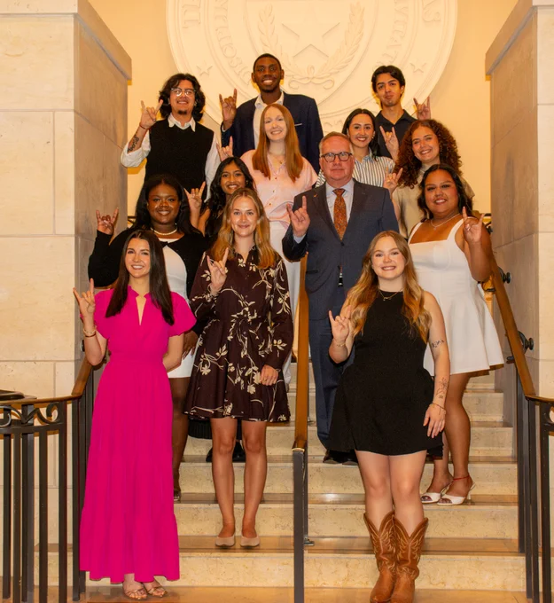 A group of people posing on a staircase, making the Hook 'em Horns hand gesture. A seal is visible on the wall behind them.