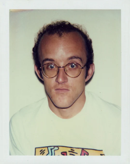 Photograph of Keith Haring wearing a white T-shirt featuring his artwork. He is looking directly at the camera.