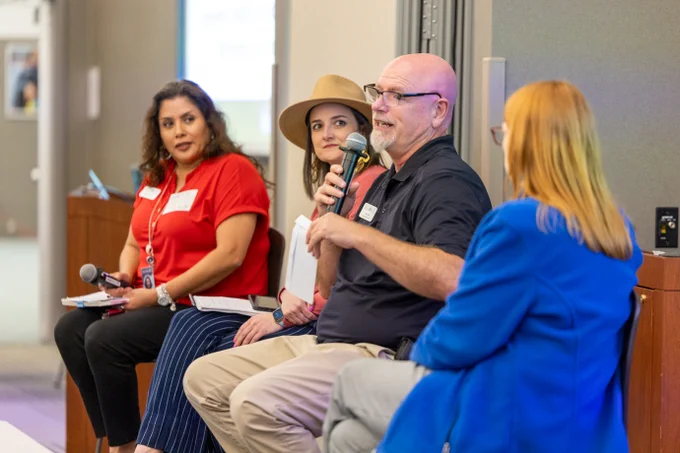 A panel of four staff members are seated, engaging in a discussion. Tom Call is holding a microphone. They are sitting in front of a screen, with conference badges visible.
