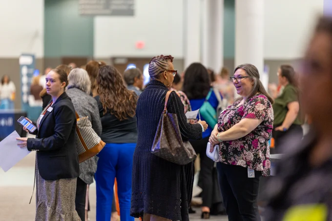 A group of individuals engaged in conversations at the Admin Advantage Conference. Various people, some holding papers, are standing in an open space with informational booths visible in the background. 