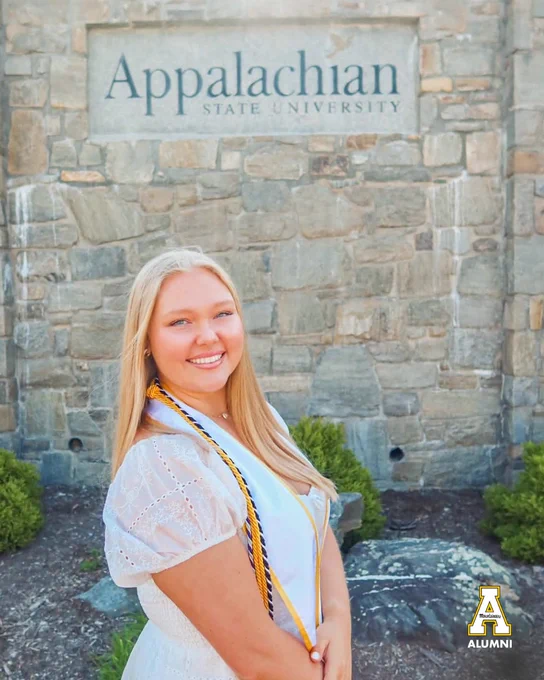Laci Hentschel smiles in a white dress and graduation stole, standing in front of a stone wall with an “Appalachian State University” sign.