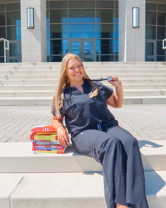Laci Hentschel sits on campus steps in black scrubs, holding a stethoscope, with a stack of textbooks beside her outside Leon Levine Hall of Health Sciences.