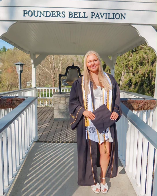Laci Hentschel stands in cap and gown at the Founders Bell Pavilion, holding her diploma cover with the campus bell visible behind her.
