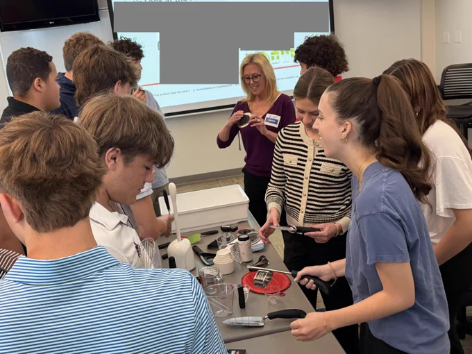 A group of students gathered around a guest speaker in a classroom, examining what appears to be kitchen & household appliances. The teacher is standing and interacting with the students, who are focused on the materials in front of them.