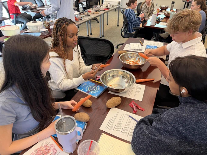 Students sitting around a table, engaged in an activity with various materials, including books, pencils, and peeling carrots & potatoes.