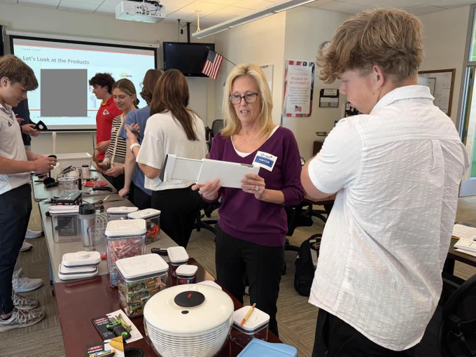 A group of people gathered around a classroom, with a woman in a purple shirt standing at the front of the room. There appears to be some kind of presentation or activity taking place.