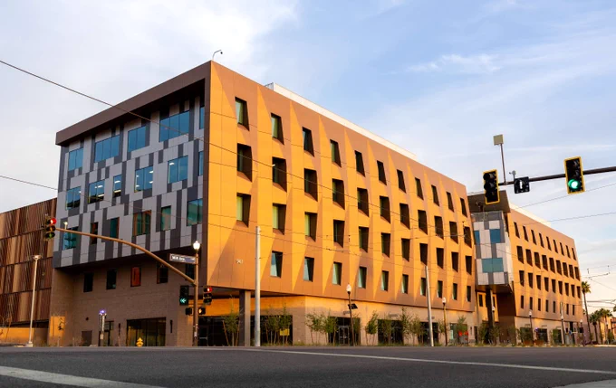 Modern multi-story building with a yellow facade and numerous windows under a partly cloudy sky at an urban intersection.