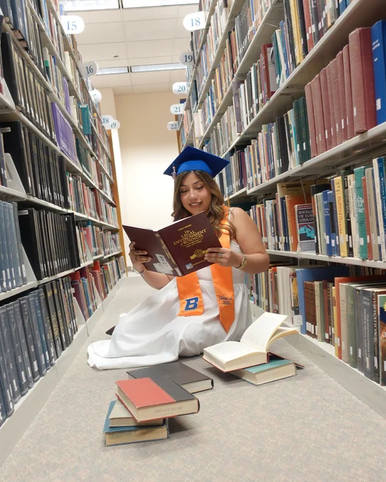 Boise State student wearing their graduation cap and stole sitting in the Albertsons Library surrounded by books. 
