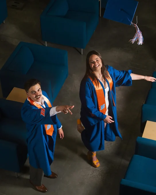 Two Boise State students standing in the Center for the Visual Arts tossing their graduation caps into the air.
