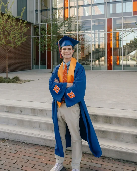Boise State engineering standing and smiling in graduation regalia in front of the Micron School of Materials Science and Engineering building. 

