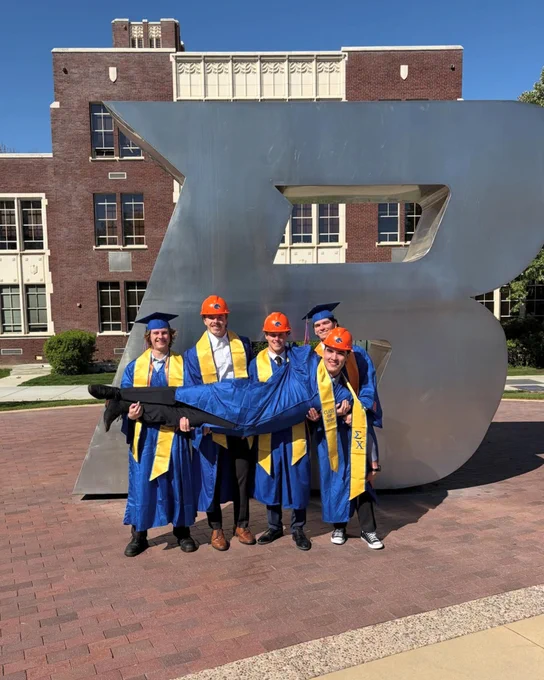 Group of Boise State students wearing graduation robes and wearing construction hats holding up their friend in the B Plaza. 
