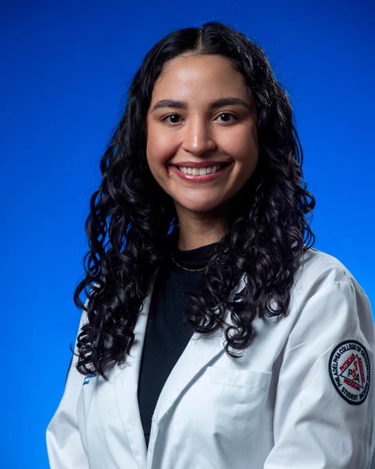 A person with curly hair is smiling against a blue background. They are wearing a white coat with a patch.