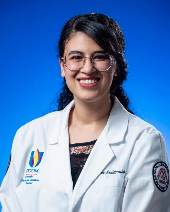 The image shows a person with long hair, wearing glasses and a white coat with the PCOM Georgia logo and a circular patch. The background is blue.