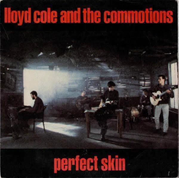 Promotional image for “Perfect Skin” (1984) by Lloyd Cole and the Commotions, featuring the band performing in a dimly lit industrial interior space. Members are positioned across the room with instruments, surrounded by shadows and soft light, creating a moody, atmospheric setting that reflects the song’s sharp, introspective tone.