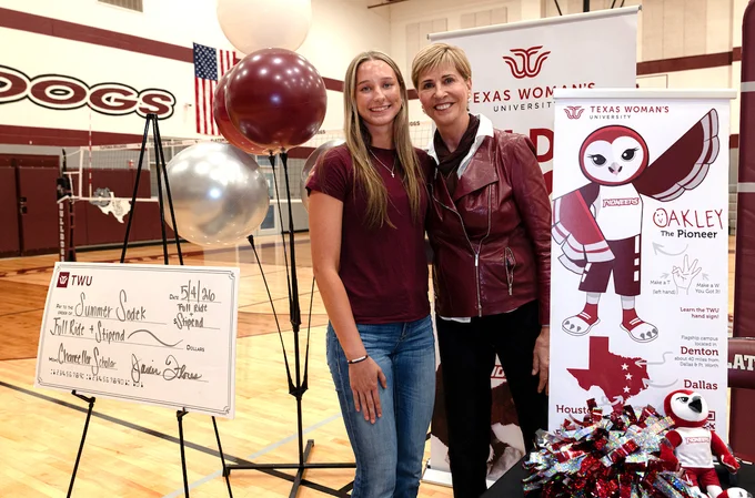 This image shows Summer Sodek and Chancellor Feyten standing in a gymnasium next to a large ceremonial check. Balloons and a Texas Woman's University backdrop with the mascot, Oakley the Pioneer, are visible. A pom-pom arrangement is on a table nearby.