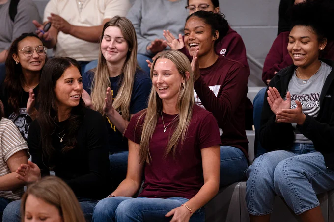 A group of people with Summer Sodek at the center, sitting in bleachers, smiling and clapping. They appear to be enjoying an event, possibly in a gymnasium setting. Some are wearing casual clothing with team or university logos.