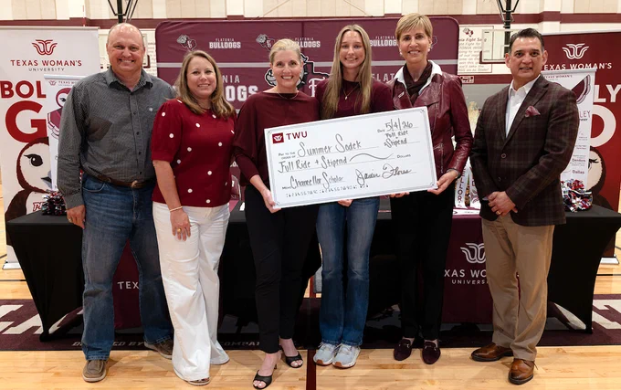 A group of people stands together in front of a backdrop featuring Texas Woman's University branding. They are smiling and holding a large, ceremonial check labeled "Summer Sodek Full Ride Scholarship." The setting appears to be an indoor event space with additional university and bulldog-themed banners.