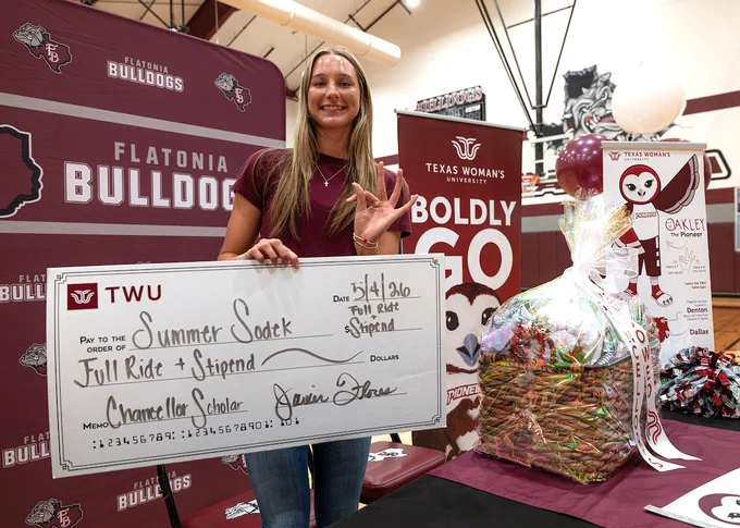 Summer Sodek is holding a large check inside a gymnasium with "Flatonia Bulldogs" banners. The check, from Texas Woman's University (TWU), is made out to Summer Sodek, marked for a full ride and stipend. A gift basket with multicolored items is nearby.