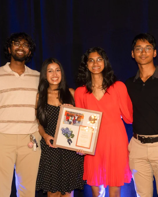 Slide 2: 4 students stand together holding a plaque and smiling.