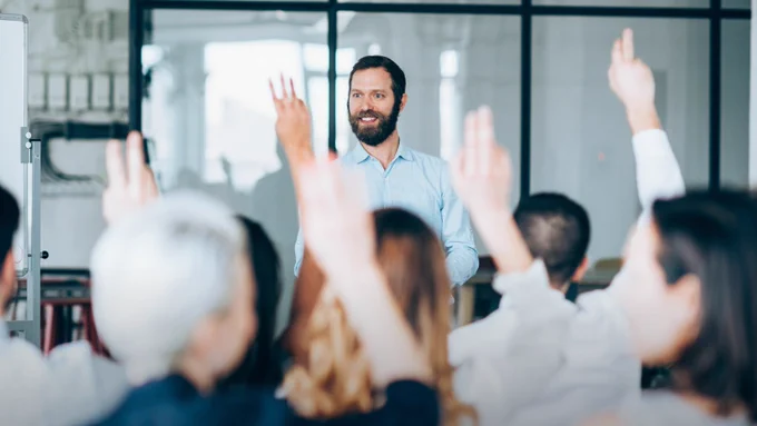 Image of a person standing at the front of a classroom, smiling, with several attendees raising their hands in a modern room.