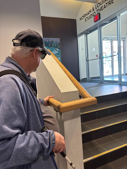 Tim Nolan walking up the stairs in a hallway of the theatre leading towards a set of doors, with the words "Norman and Louise studio theatre" above the glass doors leading to the outside. 