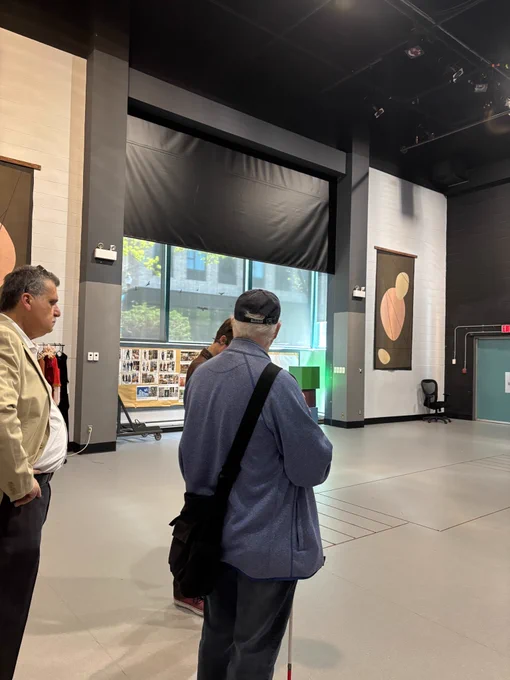 Image of theatre lobby with grey floors, black ceiling, and a window with black curtains covering the top hald. Tim Nolan is in the centre of the image, with his back facing the camera, and Dr. Samir Chidiac on the left.