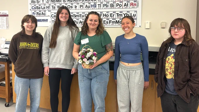 Lindsey Kiecker standing between 4 students, holding flowers. Periodic Table on the wall behind them