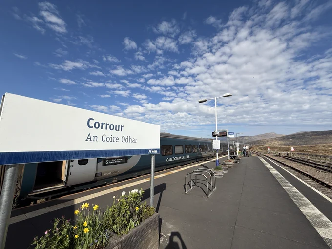 Corrour station sign with Caledonian Sleeper train in the background - the sky is blue with some clouds. Mountains can be seen in the background.
