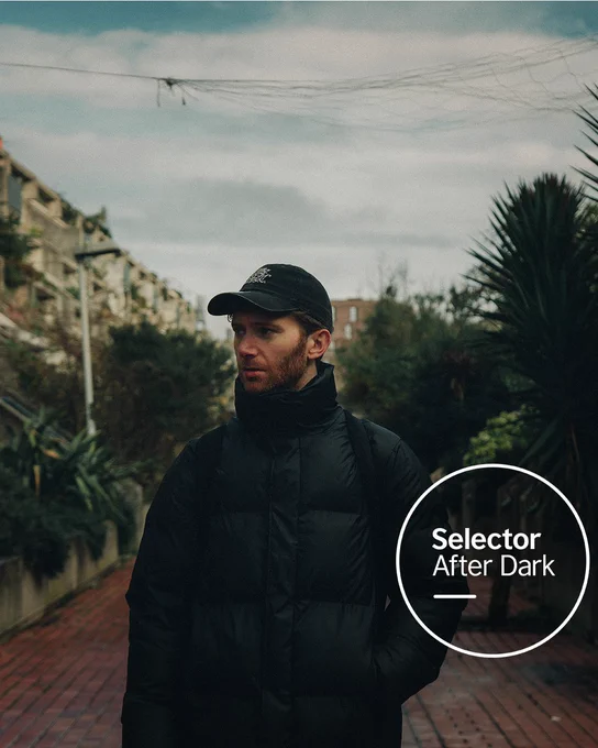 Electronic DJ and producer Fractures stands on a narrow, plant-lined urban path wearing a black cap and puffer jacket, looking off to the side under an overcast sky. The Selector After Dark logo sits in a white circle at the bottom right.