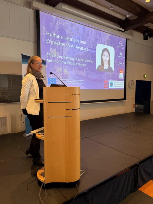 Image shows a speaker at a podium on a stage. There is a projector behind the speaker with the following text: 'Human-likeness and empathy in AI avatars. Enhancing Passenger experience in autonomous public vehicles.' The EU flag is visible on the screen, as well as the OptiPEx project logo and the IAV GmbH logo.