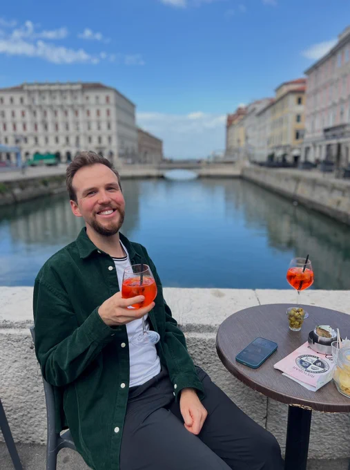 Photo of me in a white shirt with a green jacket over it, sitting in front of the Canale Grande, a water canal in Trieste, holding a glass of Aperol in my hand and smiling.