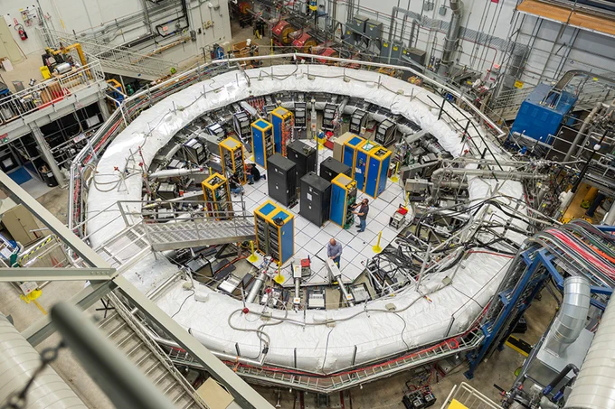 View from above of the Muon g-2 experiment, which looks like a very large ring with white plastic padding on top. There are computer cabinets and scientists standing in the middle. 