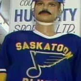 Photo of a man standing indoors wearing a blue hockey jersey with Saskatoon Blues across the front and a stylized logo. He has a thick mustache and is wearing a helmet. A sign behind him reads Humboldt Sports Ltd. The setting appears to be inside a sports or retail space with structural beams visible overhead.

