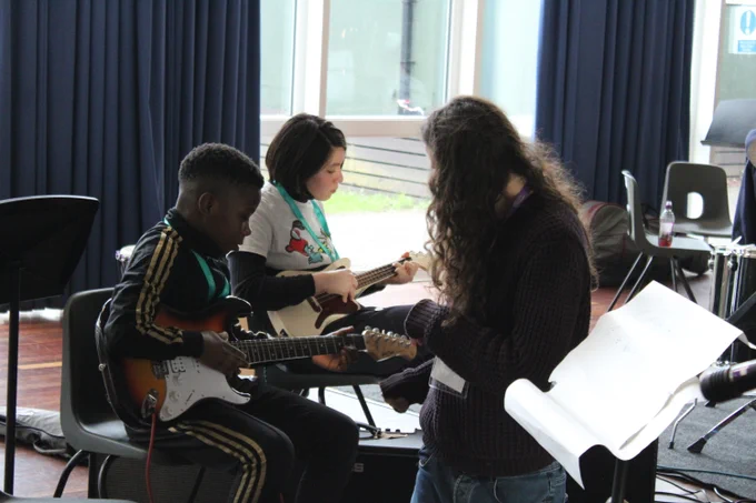 Two children seated indoors playing electric guitars while an adult with long hair instructs them during a music lesson.