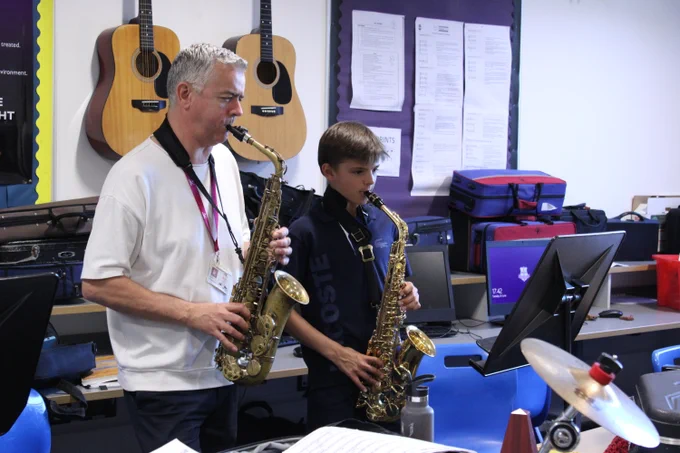 An adult and a boy playing saxophones together in a music classroom with guitars hanging on the wall behind them.