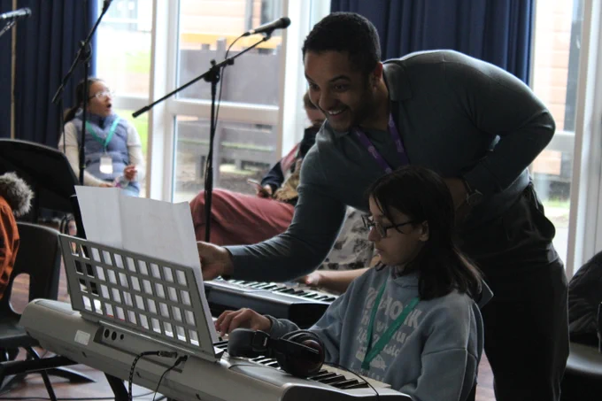 A man guides a young girl playing an electronic keyboard in a sunlit room with other seated people in the background.