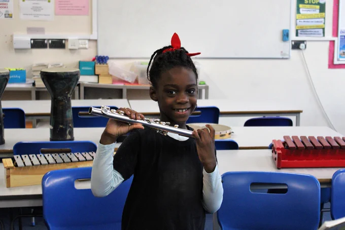 Young student practicing flute in a music classroom with xylophones and other instruments on tables behind.