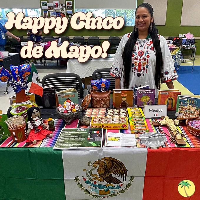Table decorated with Mexican flag and traditional snacks, crafts, and a woman in embroidered blouse celebrating Cinco de Mayo indoors.