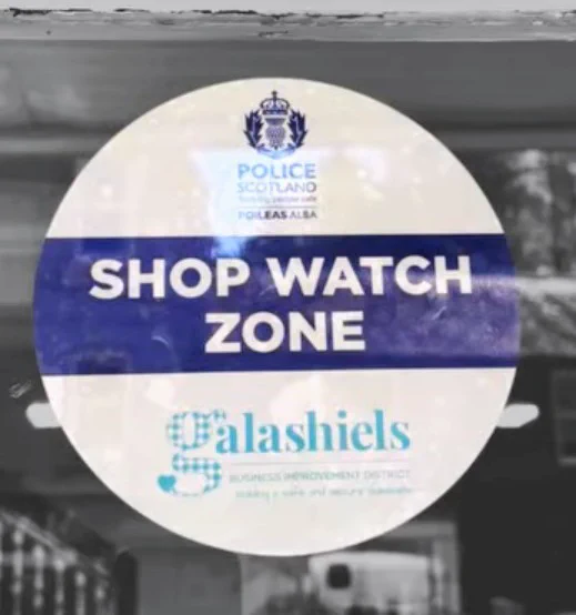 Sticker on a window. The sticker is circular in shape with a white background. It has a Police Scotland logo and Galashiels logo with Shop watch zone written on a navy banner across the middle.