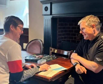 A woman and a man sit at a table discussing documents, with a fireplace in the background.
