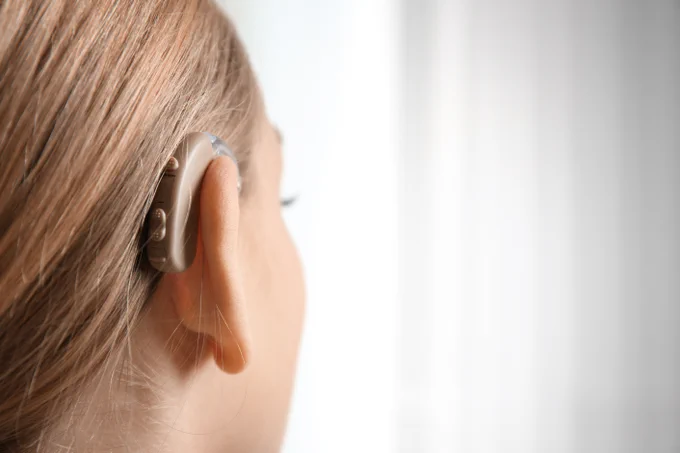 Close-up of a person wearing a modern behind-the-ear hearing aid on their left ear against a bright background.