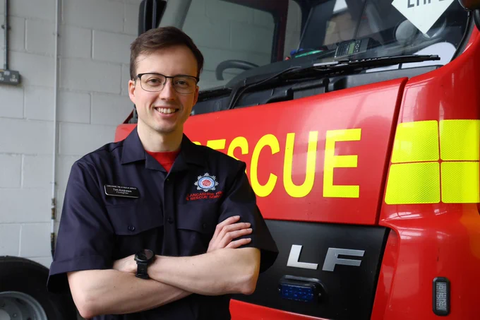 A male firefighter with short, dark hair and glasses and wearing blue duty-rig uniform standing with arms crossed in front of a red rescue fire truck inside a fire station.