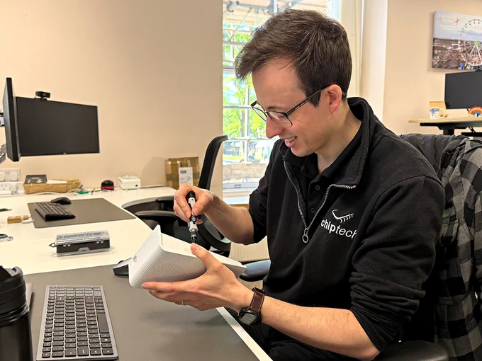 A man with dark hair and glasses holding a screwdriver and working on a white electronic device known as a SEVEN. He is sat at a desk in a well-lit office with computer equipment, and smiling as he works.
