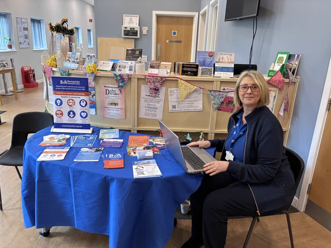 A woman from Basingstoke Voluntary Action (BVA) sat in a reception area at a table and holding a laptop, with leaflets for various services BVA provides.