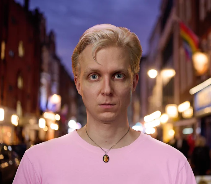 Linus Karp, a blonde man with blue eyes is looking directly into camera. He's wearing a pink t-shirt and a necklace with an image of Princess Diana. He's standing against a blurred out background of a street in Soho.
